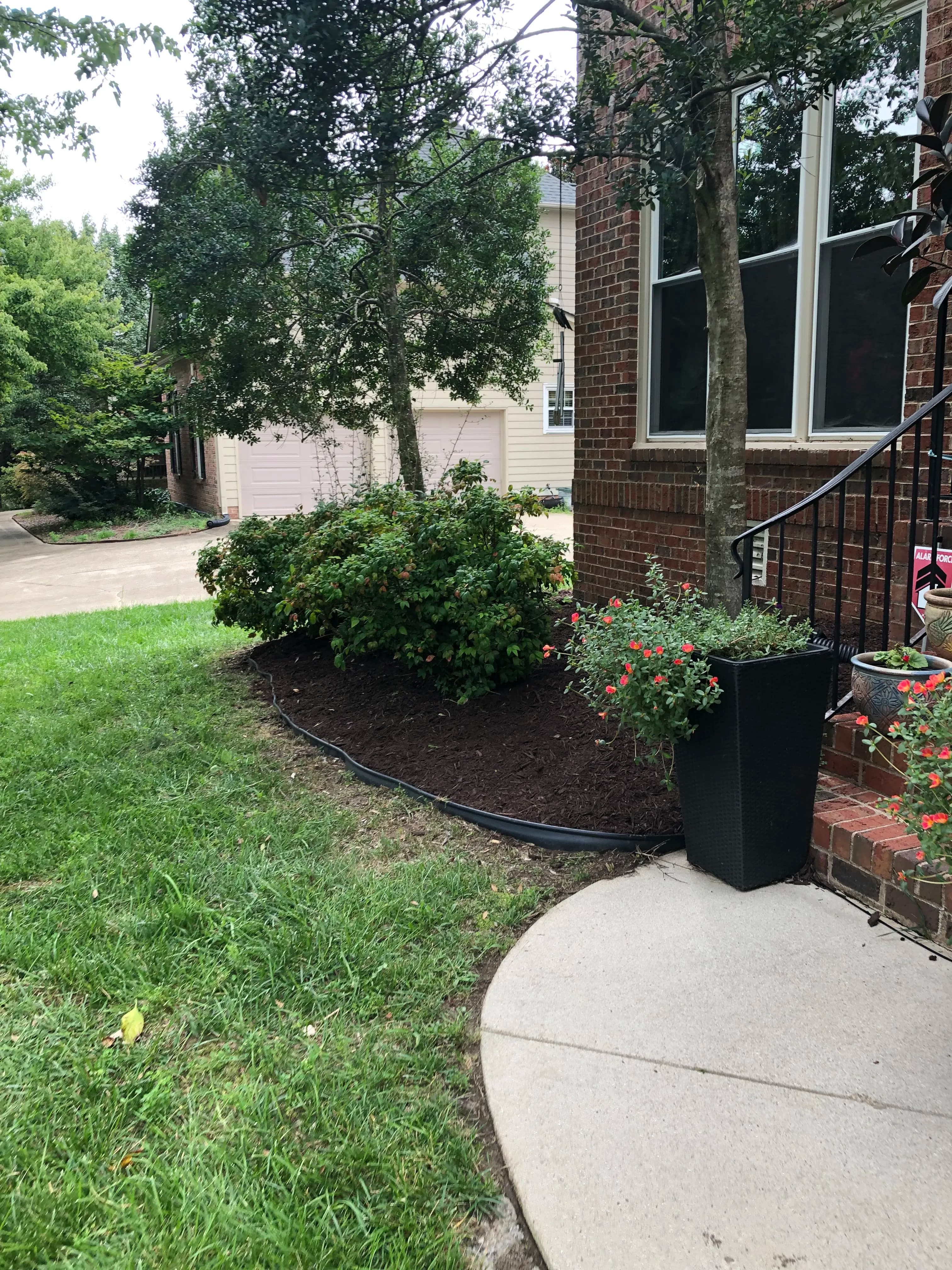 Landscape design featuring shrubs, flowers, and a concrete pathway near a brick home.